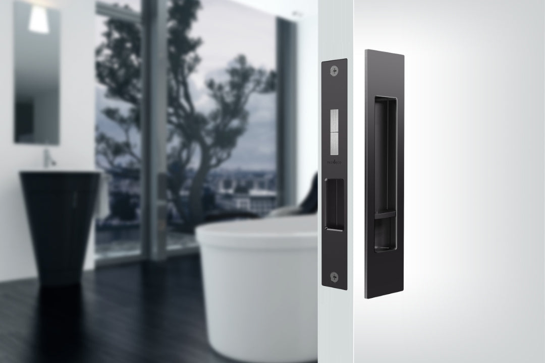 Insitu image of the BL8004/SET Matt Black Mardeco 'M' Series Cavity Sliding Door Set installed on a white door with a blurred out bathroom in the background.