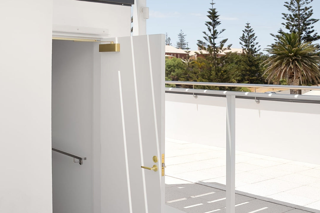 Insitu image of the Satin Brass Door Closer with Slide Arm on a white door and palm trees in the background.