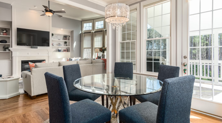 Dining room with blue chairs around a glass table. In the background are white windows and a ceiling fan.