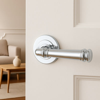 The Apollo Design Tuscany Door Handle in chrome plate installed on a cream door with a lounge room in the background.