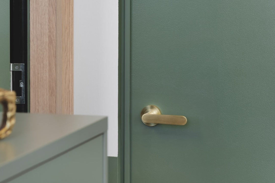 The Duke Satin Brass Door handle installed on a green door with a green cupboard in the foreground.