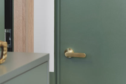 The Duke Satin Brass Door handle installed on a green door with a green cupboard in the foreground.