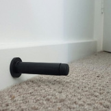 The Matt Black Wall Mount Door Stop on a white skirting and carpet for flooring.