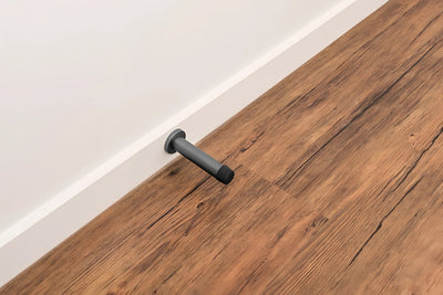 The Zanda Graphite Nickel Round Wall Door Stop 85mm installed onto the skirting board with a wooden floor in the picture as well.