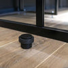 Insitu product picture of the Matt Black Round Floor Door Stop 38mm installed on a wooden floor in front of black door.