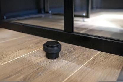 Insitu product picture of the Matt Black Round Floor Door Stop 38mm installed on a wooden floor in front of black door.