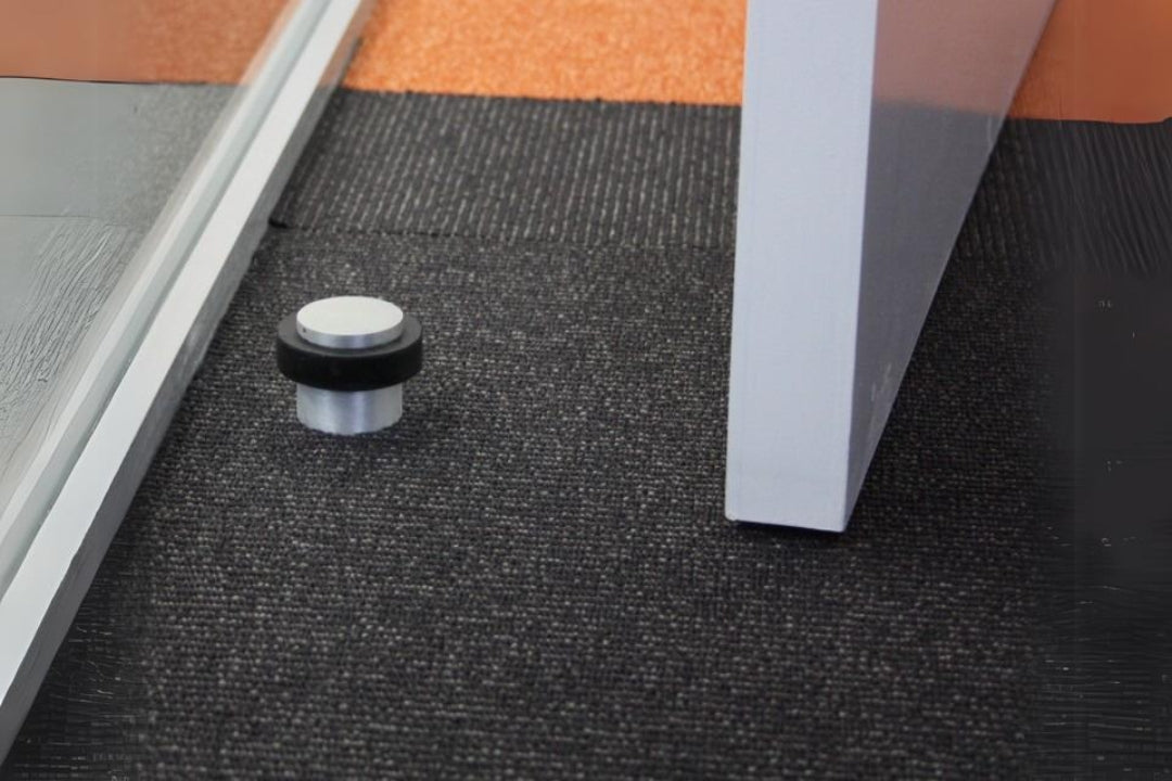 Insitu image of the Brushed Nickel Round Floor Door Stop 38mm installed on a carpet floor with a door in picture and also a glass wall.
