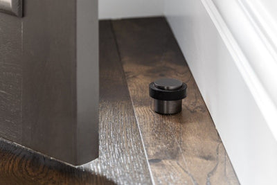 The Gun Metal Grey Round Floor Door Stop 38mm installed on brown floor boards and white walls in the picture as well as a door about to hit the door stop.