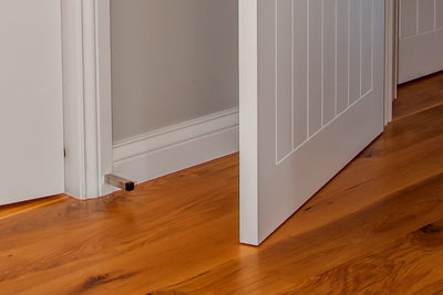 The Zanda Square Skirting Mount Door Stop 80mm Brushed Nickel 5262.BN installed on white skirting with a white door and polished wooden floors.