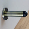 The Antique Brass Pencil Wall Mounted Door Stop installed on the wall with a wooden floor.