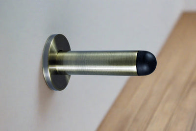 The Antique Brass Pencil Wall Mounted Door Stop installed on the wall with a wooden floor.