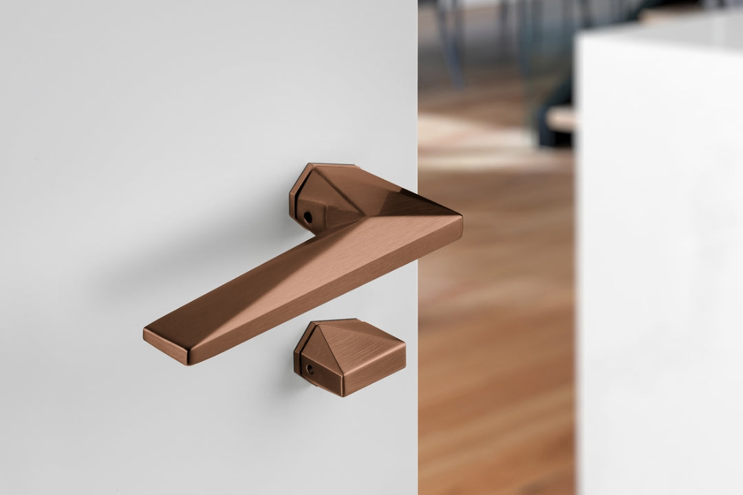 Archi Sketch Titanium Copper lever handle on a white door with a blurred wooden floor background