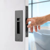 Insitu image of the IN.20.925.TB Matt Black Sliding Door Privacy Kit on an off white door. A hand is pulling the door via the edge pull with a blurred out bath in the background.