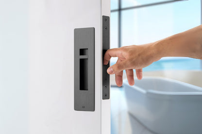 Insitu image of the IN.20.925.TB Matt Black Sliding Door Privacy Kit on an off white door. A hand is pulling the door via the edge pull with a blurred out bath in the background.