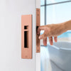 Insitu image of the IN.20.925.TCO Copper Sliding Door Privacy Kit on an off white door. A hand is pulling the door via the edge pull with a blurred out bath in the background.