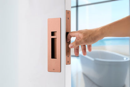 Insitu image of the IN.20.925.TCO Copper Sliding Door Privacy Kit on an off white door. A hand is pulling the door via the edge pull with a blurred out bath in the background.