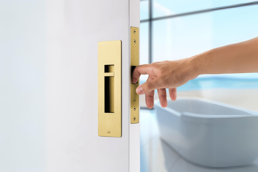 The Pendulum Sliding Door Privacy Kit in Satin Brass installed on an off white door with a bath in the background. A person's hand is pulling the door by the edge pull.