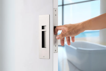 Insitu image of the IN.20.925 Satin Stainless Steel Sliding Door Privacy Kit on an off white door. A hand is pulling the door via the edge pull with a blurred out bath in the background.
