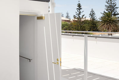Insitu image of the Satin Brass Door Closer with Slide Arm on a white door and palm trees in the background.
