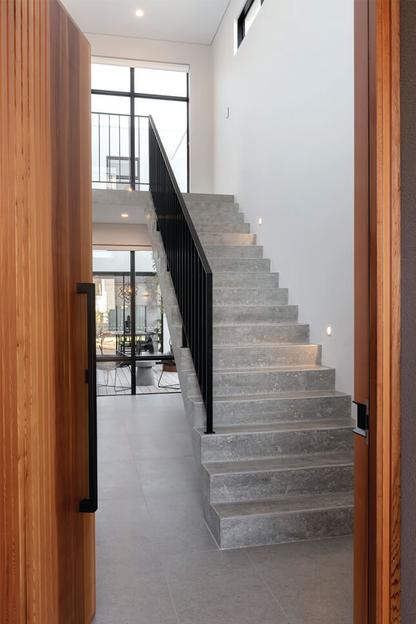 A matt black Ultra pull handle installed on a wooden door with a view inside where there is a concrete staircase and black railing.